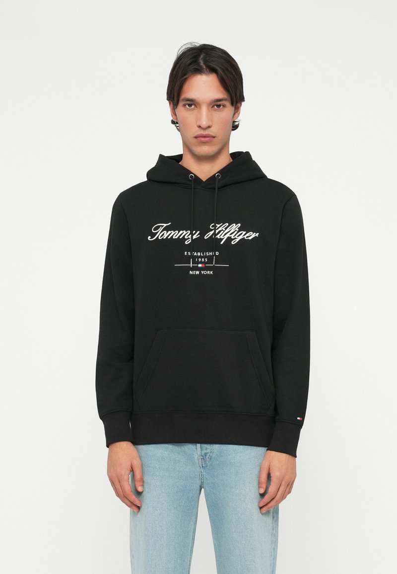 Hoodie noir en coton avec un cordon de serrage. Présente un texte logo blanc sur le devant, une poche kangourou, et des poignets et un ourlet côtelés.