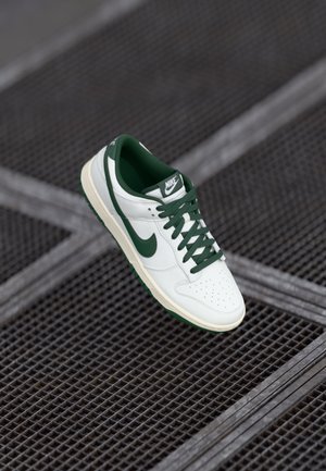 White and green Nike sneaker floating above a metal grate floor, showing side and top views of the shoe.