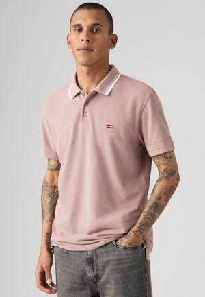 Jeune homme aux cheveux courts et aux bras tatoués porte un polo Levi's rose clair et un jean gris, debout devant un fond uni.