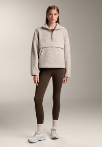 Fleece pullover in lichtbeige met een halve rits en een oversized kraag, gecombineerd met bruine leggings en lichtgrijze sneakers. Zachte textuur zichtbaar.