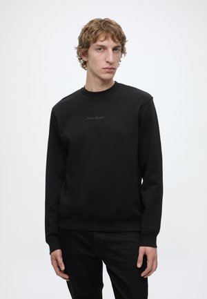 Jeune homme aux cheveux bouclés portant un sweat-shirt noir et un pantalon noir, debout devant un fond gris clair uni.