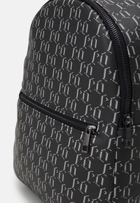 Ryggsäck i svart material med ett upprepande grått logomönster. Anmärkningsvärda detaljer inkluderar en ficka med dragkedja fram och en slät textur.