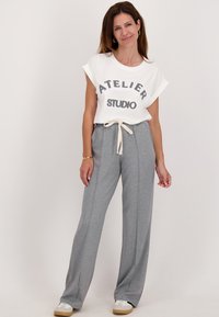 Vrouw die glimlacht, gekleed in een witte T-shirt met opgerolde mouwen met de tekst "Atelier Studio", grijze broek met koordjes, witte sneakers en gouden creolen.