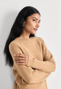 Femme aux longs cheveux noirs portant des boucles d'oreilles dorées et un pull en tricot beige, les bras croisés, regardant sur le côté devant un fond uni.