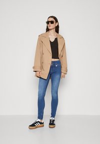 Bežna oversize trench jakna, črn usnjen top, modri skinny jeans in črne teniske z belimi in gumastimi detajli; sončna očala zaokrožajo videz.