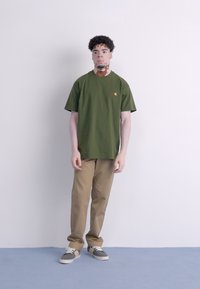 Grøn bomulds T-shirt med korte ærmer og rund halsudskæring, der har et lille logo. Sammen med beige bukser og grå sneakers.