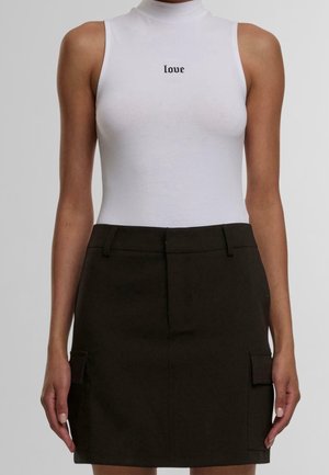 Kvinde iført en hvid ærmeløs top med teksten "love" og en sort mininederdel med sidelommer på en neutral baggrund.