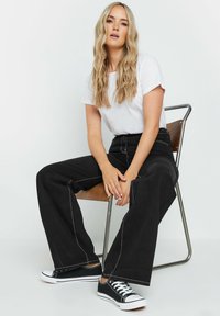 Pantalones de pierna ancha negros con costuras blancas en contraste, combinados con una camiseta blanca sencilla y zapatillas negras, sentado en una silla de metal y madera.