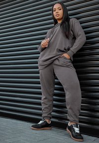 Grå ribbad hoodie med framficka, ihopparad med matchande byxor. Svarta sneakers med vita snören och en gummisul. Bakgrunden har en svart jalusi.