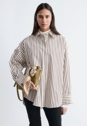 Frau trägt ein übergroßes weißes Hemd mit braunen Streifen, hält eine olivgrüne Handtasche mit einem gemusterten Schal und trägt schwarze Hosen.