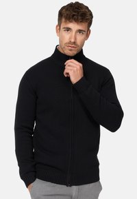 Pull noir à col haut zippé avec des côtes. Comprend une fermeture éclair à l'avant et des poignets ajustés, fabriqué en matière tricotée.