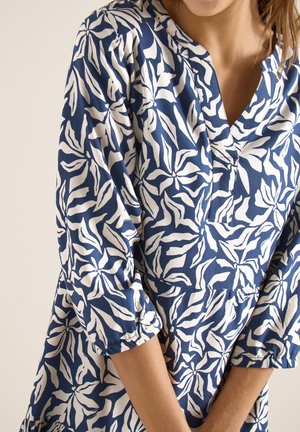 Frau trägt eine blau-weiße Bluse mit Blumenmuster, drei Viertel langen Ärmeln und V-Ausschnitt.