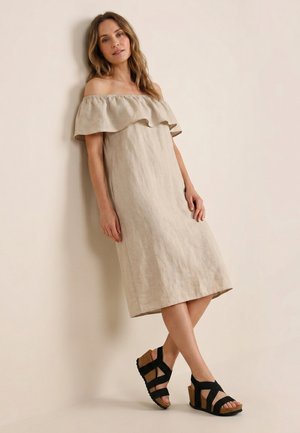 Kvinde lænet op ad væg iført en beige off-shoulder midi-kjole og sorte kilesandaler med korksåler.