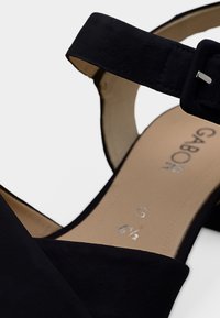 Μαύρο σανδάλι από σουέντ με λουράκι στον αστράγαλο και ασημένια αγκράφα, ανοιχτή μύτη, μαλακό πέλμα, με ετικέτα που αναγράφει το μέγεθος και το λογότυπο της μάρκας.