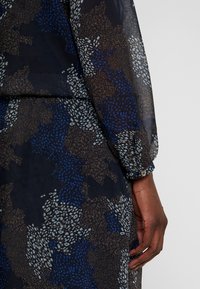 Průsvitné šaty s vícetrobným potiskem v námořnické modré, hnědé a modré. Mají nařasené rukávy s detailem knoflíků. Měkká, lehká látka.