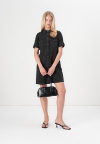 Zwarte spijkerstof shirtjurk met korte opgerolde mouwen, knoopsluiting aan de voorkant, twee borstzakken, gecombineerd met een kleine zwarte handtas en zwarte slippers.