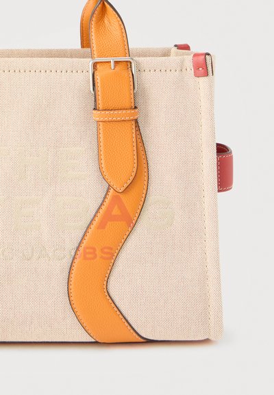 Μπεζ καμβά τσάντα tote με πορτοκαλί δερμάτινο λουρί που διαθέτει αγκράφα και κόκκινη δερμάτινη πλαϊνή ετικέτα, με εντυπωμένο κείμενο που φαίνεται μερικώς στο μπροστινό μέρος.