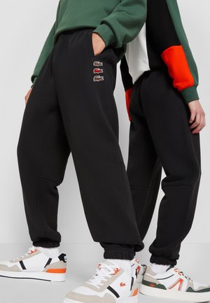 Deux personnes portant des pantalons de survêtement noirs avec des logos de crocodile brodés, associés à des baskets blanches et à des sweats à capuche bicolores vert et rouge.