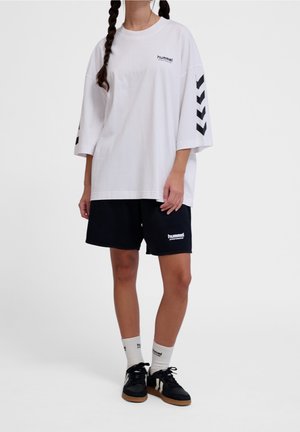 Persona que lleva una camiseta blanca extragrande de Hummel con franjas en forma de cheurón negras en las mangas, pantalones cortos negros de Hummel, calcetines blancos y zapatillas negras.