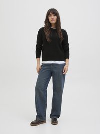 Μαύρος πλεκτός σγουρός με ύφανση ραβδώσεων, φορεμένος πάνω από λευκό πουκάμισο, σε συνδυασμό με χαλαρούς μπλε τζιν και σκούρα παπούτσια με παχύ πάτο.