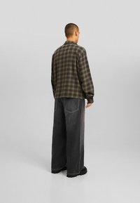 Camisa de cuadros extragrande en verde y negro, hecha de un tejido texturizado; combinada con pantalones de denim de pierna ancha en gris oscuro.