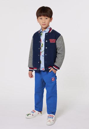 Veste universitaire bleu marine et grise avec boutons sur le devant, accents rouges ; chemise à motifs bleu clair ; pantalon bleu vif ; chaussures blanches brodées avec des détails.