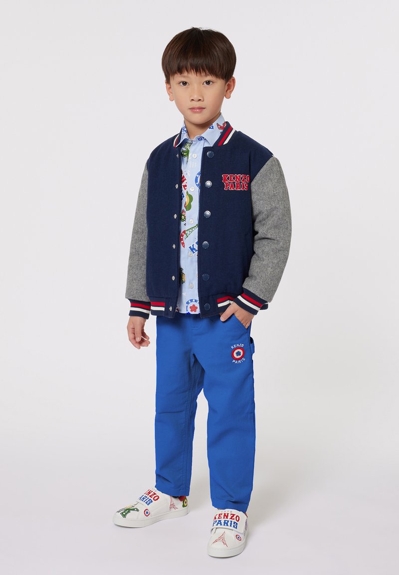 Marineblauwe en grijze varsityjack met knoopsluiting, rode accenten; lichtblauw patroonoverhemd; felblauwe broek; geborduurde witte schoenen met details.