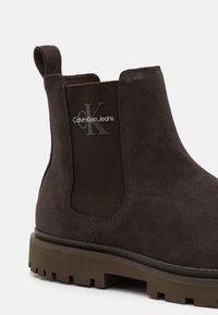 Botte de cheville en daim marron avec des panneaux latéraux élastiques, signée "Calvin Klein Jeans", et avec une semelle extérieure robuste marron.