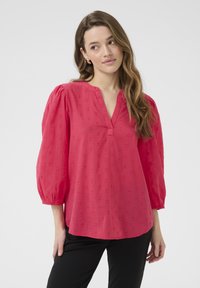 Blouse rose vif avec un décolleté en V, des manches bouffantes, un motif de cœurs brodé et une coupe décontractée, associée à un pantalon noir.