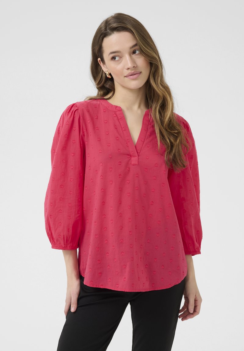 Blouse rose vif avec un décolleté en V, des manches bouffantes, un motif de cœurs brodé et une coupe décontractée, associée à un pantalon noir.