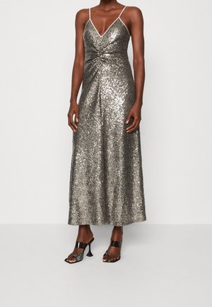 Mujer con vestido midi sin mangas plateado de lentejuelas con tirantes finos y detalle de torsión, combinado con sandalias negras de tacón alto y punta abierta.