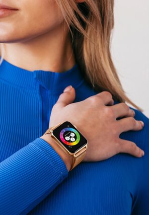 Montre intelligente avec un bracelet en maille dorée, écran carré affichant une interface colorée, portée sur un poignet avec un t-shirt de sport bleu texturé.
