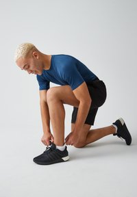 Zwarte sport schoenen met een gestructureerde bovenkant en witte zool, gecombineerd met een blauwe strakke shirt en zwarte shorts. Persoon knielt om de schoenen te strikken.