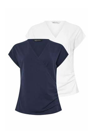 Blonde vrouw die glimlacht, gekleed in een wit T-shirt met V-hals en korte mouwen, en zwarte broek, staand met één hand op de heup tegen een witte achtergrond.