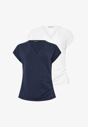 Blonde vrouw die glimlacht, gekleed in een wit T-shirt met V-hals en korte mouwen, en zwarte broek, staand met één hand op de heup tegen een witte achtergrond.