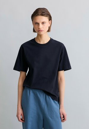 Jong persoon met kort bruin haar die een zwarte T-shirt met korte mouwen en losse blauwe broek draagt tegen een effen grijze achtergrond.