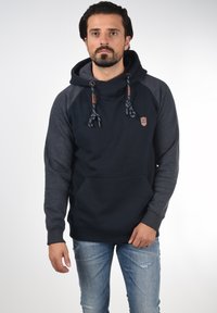 Sudadera azul oscura con mangas raglán grises, presenta un bolsillo canguro, cordones con detalles estampados y un parche de logo en el pecho.
