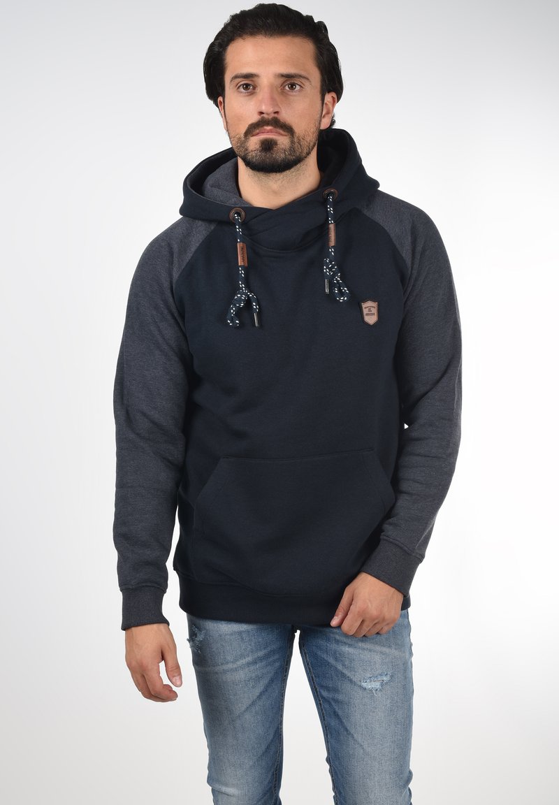 Sudadera azul oscura con mangas raglán grises, presenta un bolsillo canguro, cordones con detalles estampados y un parche de logo en el pecho.
