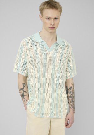 Jeune homme tatoué portant un polo à manches courtes en maille ouverte rayé bleu clair et crème, et un pantalon crème, debout devant un fond uni.