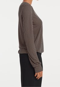 Femme portant un chemisier à manches longues à carreaux marron et blanc, associé à un pantalon noir, vue de profil sur un fond uni.