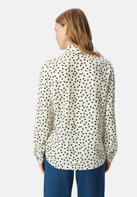 Blusa a maniche lunghe in tessuto crema con pois neri, dotata di colletto e orlo curvo, indossata con pantaloni in denim blu.
