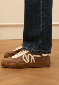 Bruine suède sneakers met witte accenten, platte veters en een rubberen zool. Gecombineerd met donkere denim jeans, waardoor het ontwerp en de textuur van de schoenen zichtbaar zijn.