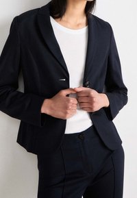 Blazer bleu marine à revers crantés et boutons doubles, associé à une chemise blanche. Coupe ajustée avec des épaules structurées et une texture de tissu lisse.