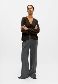 Gilet en tricot marron avec un col en V et un détail à nouer, associé à un pantalon gris ample en tissu texturé.
