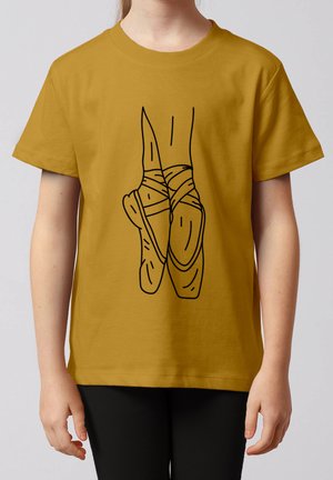 Kind trägt ein senfgelbes T-Shirt mit einer schwarzen Linienzeichnung von gekreuzten Ballett-Spitzenschuhen auf der Vorderseite.