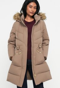 Mujer con un abrigo largo acolchado beige con capucha de piel sintética, detalles en dorado en el cierre y botones, blusa burdeos y jeans oscuros, de pie.