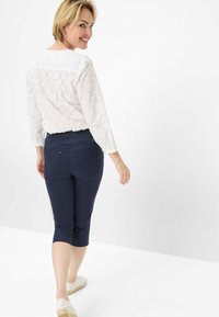Blusa bianca ricamata con scollo rotondo, abbinata a pantaloni navy a tre quarti. Il modello indossa scarpe chiare e volge le spalle alla macchina fotografica.