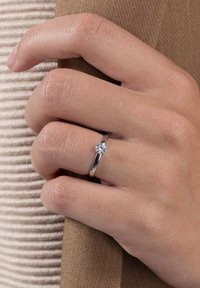 Silberner Ring mit einem runden, klaren Stein, ausgestattet mit einem schmalen Band und einer Vierkrallenfassung, getragen an einer Hand vor einem strukturierten Stoffhintergrund.