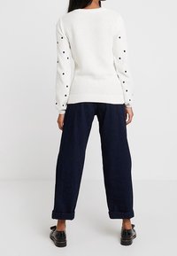 Frau in einem weißen langärmeligen Pullover mit schwarzen Punkten auf den Ärmeln, blauen weiten Hosen mit Umschlag und schwarzen Schuhen, von hinten gesehen.