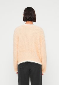 Πουλόβερ πλεκτό ροδακινί με χνουδωτή υφή, λευκή επένδυση στο τελείωμα και μακριά μανίκια. Φορεμένο με σκούρο γκρι παντελόνι, εμφανισμένο από την πίσω πλευρά.